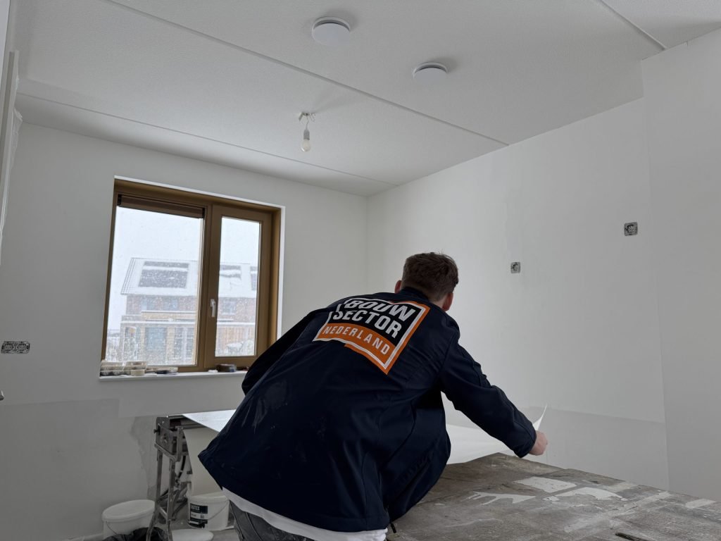 Vakman van Bouwsector Nederland strijkt een baan behang glad op de plaktafel bij het raam